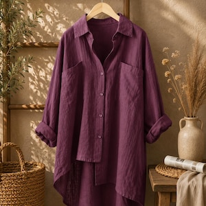 May include: A deep purple long-sleeve button-down shirt featuring a collared neckline, two large chest pockets, and an asymmetrical hemline that is shorter in the front and longer in the back, displayed on a wooden hanger.