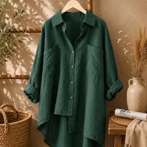 May include: A dark green button-down shirt with a collar, long sleeves with rolled cuffs, and two chest pockets. The shirt features an asymmetrical hemline that is shorter in the front and longer in the back, crafted from a textured fabric.