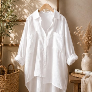 May include: A crisp white button-down shirt featuring a classic collar, long sleeves with rolled cuffs, and two front chest pockets. The garment has a high-low hemline with a longer back panel and is displayed on a wooden hanger.