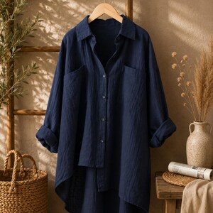 May include: A dark navy blue long-sleeve button-down shirt with a collared neckline, two front chest pockets, and a high-low hemline. The shirt features a textured fabric finish and is displayed on a wooden hanger with sleeves rolled up.