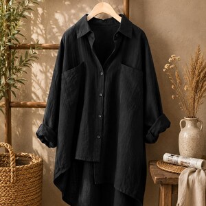 May include: A black long-sleeved button-down shirt with a collar, two front chest pockets, and a high-low hemline. The shirt is made of a textured fabric and is displayed on a wooden hanger, featuring rolled-up sleeves and a buttoned front placket.