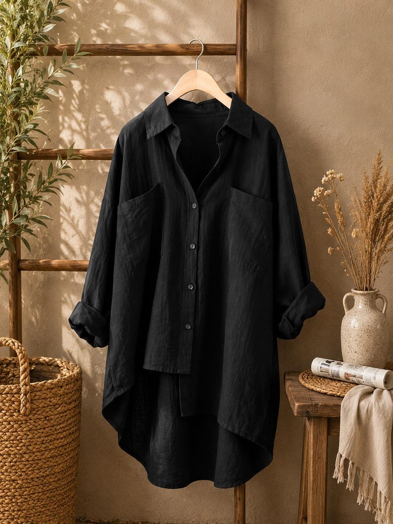 May include: A black long-sleeved button-down shirt with a collar, two front chest pockets, and a high-low hemline. The shirt is made of a textured fabric and is displayed on a wooden hanger, featuring rolled-up sleeves and a buttoned front placket.