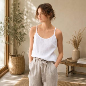 May include: A white sleeveless camisole top with thin shoulder straps and a scoop neckline, paired with high-waisted beige linen trousers featuring a matching fabric tie belt at the waist and a relaxed, straight-leg fit.
