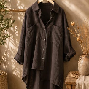 May include: A dark brown long-sleeve button-down shirt with a collar, two front chest pockets, and a high-low hemline. The shirt features a textured fabric, cuffed sleeves, and a button-up front closure, hanging on a wooden hanger.