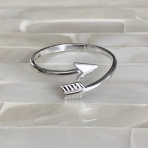 May include: A silver ring with a simple arrow design. The arrow is pointing to the right and is wrapped around the band of the ring.