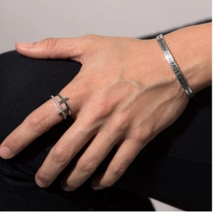 May include: A silver ring with a double cross design, set with small clear stones. The ring is worn on the index finger of a hand also wearing a silver bracelet with the inscription "Courage, Strength, and Wisdom".
