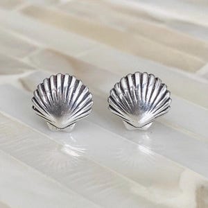 May include: A pair of silver-toned stud earrings shaped like scallop shells. Each earring features a detailed, fan-like ribbed texture radiating from the base to the rounded edge, capturing the natural form of a seashell in a metallic finish.
