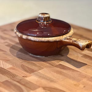 May include: A round, brown ceramic soup bowl with a matching lid and a single side handle. The bowl features a glossy dark brown glaze with a textured, lighter beige drip pattern along the rim of the lid, the top edge of the bowl, and the handle.