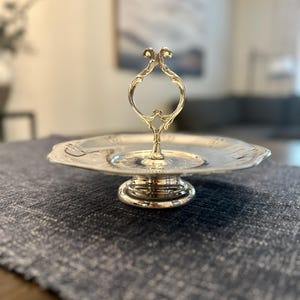Peut inclure: Un plateau de service en métal argenté avec une base circulaire et une poignée verticale centrale. Le plateau présente une assiette peu profonde et large avec un bord festonné et une poignée ajourée ornée qui s'élève du centre du plat.