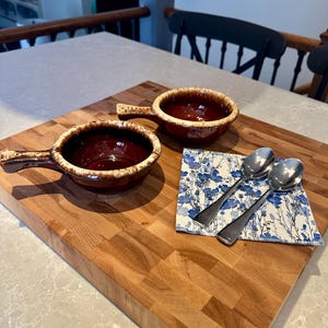 May include: Two round ceramic bowls with handles, featuring a deep brown interior and a cream-coloured drip glaze along the rim. Beside the bowls lies a square paper napkin with a blue floral pattern and two polished stainless steel spoons.
