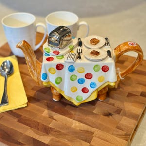 May include: A ceramic teapot shaped like a table with a white tablecloth featuring colourful polka dots. The top displays a silver toaster, two plates with toast, and cutlery. The handle features a small British flag decal, and the spout is brown.