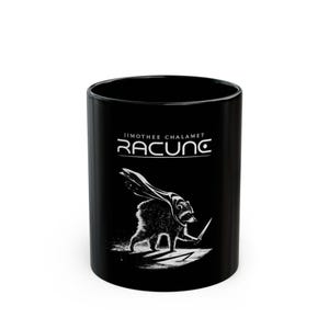 May include: A black ceramic mug featuring a white graphic illustration of a raccoon wearing a cape and holding a small blade. Above the raccoon, the text reads JIMOTHEE CHALAMET RACUNC in a stylized white font against the solid black background.