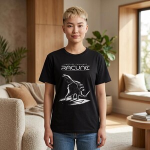 May include: A black short-sleeve t-shirt featuring a white graphic print of a raccoon wearing a cape and holding a sword. Above the graphic, the text reads "JIMOTHEE CHALAMET" and "RACUNC" in a stylized white font. The shirt has a crew neckline.