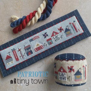 May include: A rectangular fabric piece and a round pincushion featuring cross-stitch designs of houses, pinwheels, and the text "free" and "Liberty". The items are accented with red, white, and blue twisted yarn and dark blue fabric.