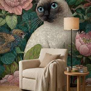 May include: A cream-coloured armchair with a matching throw blanket sits before a wall mural featuring a Siamese cat with blue eyes, large pink flowers, and birds. A small wooden side table holds a mug and a stack of books under a floor lamp.
