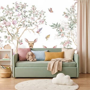 Può includere: Un divano verde salvia con cuscini rosa, blu e senape e una coperta beige. A sinistra si trova una libreria bianca con libri e un cesto intrecciato. La parete presenta un murale con alberi, fiori, farfalle e la testa di un cervo.