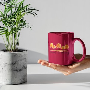 Puede incluir: Una taza de cerámica magenta con una ilustración estilo acuarela de cuatro árboles naranjas y el texto en cursiva negra "Jesus is coming" impreso en el frente. La taza tiene un asa curva y un acabado exterior liso y brillante.