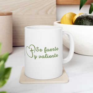 Puede incluir: Una taza de cerámica blanca con un gráfico verde de un corazón con una cruz en su interior, acompañado del texto "Se fuerte y valiente" en una fuente cursiva verde. La taza tiene un asa estándar y un diseño de cuerpo cilíndrico.