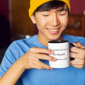 Puede incluir: Una taza de cerámica blanca con interior y asa negros, que presenta el texto impreso en negro: I wanted to do more, but Elizabeth didn't let me work. La taza tiene una forma cilíndrica y un acabado liso para contener bebidas.