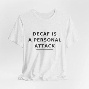 May include: A white short-sleeved t-shirt featuring the black text DECAF IS A PERSONAL ATTACK printed in a clean sans-serif font across the chest, with a horizontal line positioned directly underneath the word ATTACK on the front of the garment.