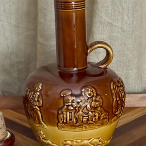 May include: A brown ceramic jug with a tall cylindrical neck and a single side handle. The body features embossed relief scenes of figures and animals in a lighter tan tone at the base, with a glossy dark brown glaze on the upper section.