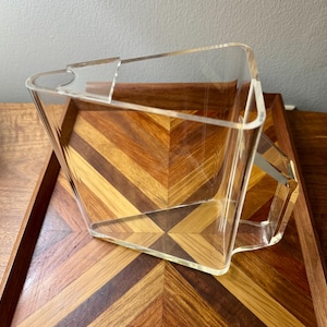May include: A clear, transparent acrylic pitcher with a geometric, angular design. The vessel features a wide, open top with a distinct pouring spout and a sturdy, integrated handle on the side, showcasing a sharp, modern, and minimalist structural form.