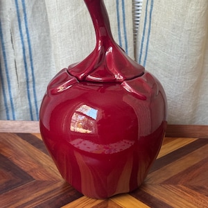 May include: A glossy, deep red ceramic jar shaped like a fruit, featuring a removable lid with a sculpted stem handle. The surface has a smooth, reflective finish with subtle indentations around the top, mimicking the natural form of an apple.