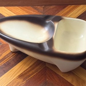 May include: A ceramic dish featuring an elongated, teardrop-shaped bowl section and a separate rectangular compartment. The piece has a cream-colored base with a dark brown, airbrushed gradient along the edges and is supported by small feet.