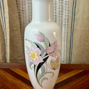 May include: A white ceramic vase with a flared rim and a tapered body. The front features a decorative illustration of pink and yellow flowers with green leaves, black accents, and a yellow and white butterfly with black details.