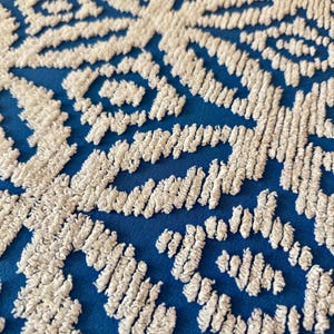 Könnte beinhalten: Ein leuchtend blauer Stoff mit einem erhabenen, weißen, strukturierten Stickmuster. Das Design besteht aus sich wiederholenden geometrischen Formen und komplexen Linien, die einen taktilen, dreidimensionalen Effekt auf der glatten, einfarbigen blauen Oberfläche erzeugen.