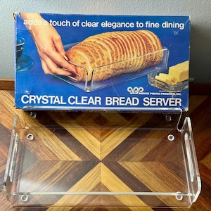 May include: A clear acrylic bread server with a rectangular base and two side handles. The product box features the text "adds a touch of clear elegance to fine dining" and "CRYSTAL CLEAR BREAD SERVER" alongside an image of sliced bread.