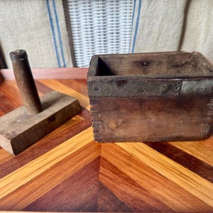 May include: A rectangular wooden butter mold with a separate wooden plunger handle. The box features finger-jointed corners and a metal band along the top edge. The wood has a dark, weathered finish with visible grain and signs of age and use.