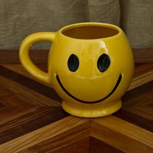 May include: A bright yellow ceramic mug featuring a black painted smiley face with two oval eyes and a wide, curved mouth. The mug has a rounded, bulbous body, a sturdy handle on the left side, and a raised circular base for stability.