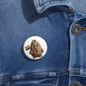 May include: A round pinback button featuring a textured, light brown, almond-shaped character with large eyes, a small nose ring, and a surprised expression. The character is holding a small, rectangular rainbow flag on a thin wooden pole.