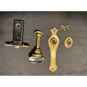 Puede incluir: Un juego de herrajes para puerta que incluye un mecanismo de pestillo de metal negro, un pomo de puerta de cristal con vástago de latón, una placa de latón alargada con grabados decorativos, un escudo circular pequeño y cuatro tornillos.