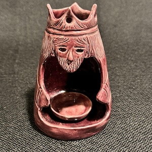 May include: A dark reddish-purple ceramic incense burner shaped like a crowned figure. The figure features a carved face with a beard and a crown at the top. A small circular bowl is integrated into the center of the hollowed-out base area.