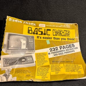 May include: A yellow and black Radio Shack BASIC Computer Language manual with a spiral binding. The cover features a vintage computer illustration, the text 232 PAGES OF PROGRAMMED INSTRUCTION AND USER PROGRAMS, and various testimonials.