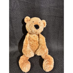 May include: A plush teddy bear with light brown, textured fur, featuring a soft, rounded head, small ears, and a prominent black nose. The stuffed animal has long, dangling arms and legs, with a simple, neutral expression and a fuzzy, soft-touch exterior.
