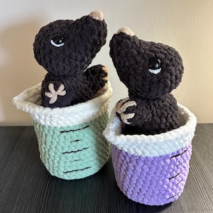 May include: Two crocheted plush moles with dark brown fuzzy bodies, beige noses, and black plastic eyes. Each mole sits inside a crocheted cup-shaped container with a white rim, one in light green and one in purple, both featuring black markings.