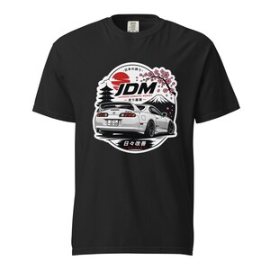 Könnte beinhalten: Ein schwarzes Kurzarm-T-Shirt mit einem Grafikdruck eines weißen Sportwagens. Das Design zeigt eine rote Sonne, eine Pagode, Kirschblüten, den Berg Fuji sowie den Text JDM, JAPANESE DOMESTIC MARKET und KAIZEN mit japanischen Schriftzeichen.