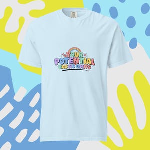Puede incluir: Una camiseta de manga corta de color azul claro con un gráfico colorido en el pecho. El diseño incluye un pequeño arcoíris y el texto "YOUR POTENTIAL HAS NO LIMITS" en letras multicolores estilo burbuja, decorado con pequeñas estrellas y corazones.