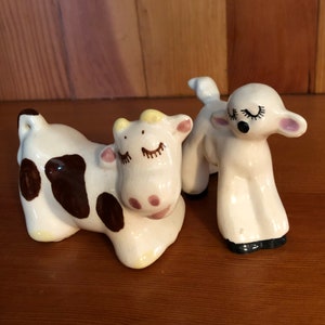 May include: A white ceramic cow with brown spots and a white ceramic lamb with closed eyes. Both figurines have painted eyelashes and pink noses.