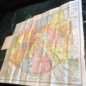May include: A vintage, folded map of Paris, France, with a detailed layout of the city's streets and neighborhoods. The map is printed in shades of yellow, orange, pink, and green, with a blue river running through the center. The title "PLAN DE PARIS" is printed in the upper left corner.