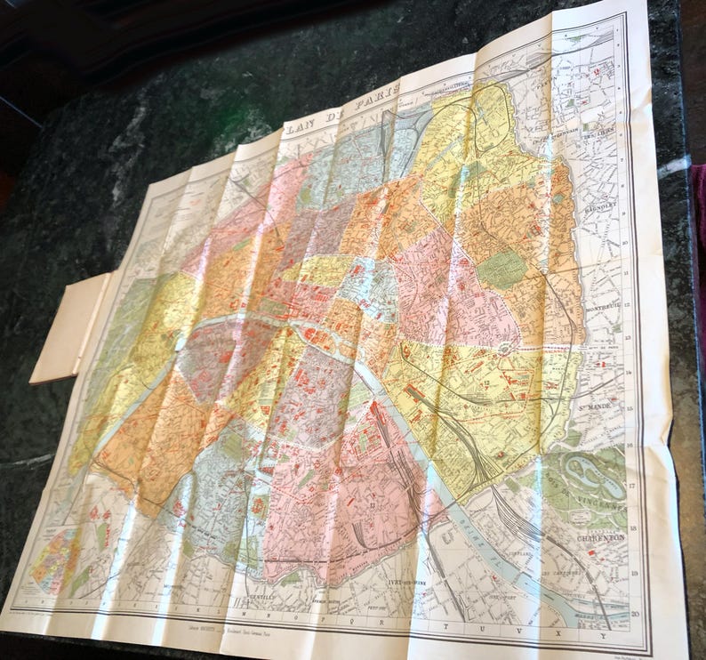 May include: A vintage, folded map of Paris, France, with a detailed layout of the city's streets and neighborhoods. The map is printed in shades of yellow, orange, pink, and green, with a blue river running through the center. The title "PLAN DE PARIS" is printed in the upper left corner.