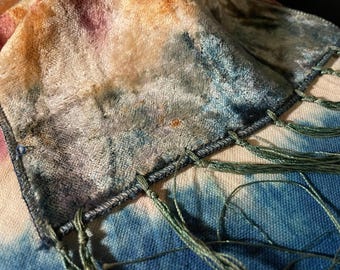 May include: A close-up of a soft, textured scarf with a tie-dye effect. The scarf features a blend of colors, including blue, green, orange, and pink. The edges are finished with a dark gray stitching and green fringe.