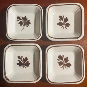 May include: Four square, off-white ceramic dishes with a brown floral design in the center. Each dish has a brown border. The dishes are arranged on a wooden surface.
