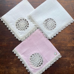 May include: Three handkerchiefs, two white and one pink, are displayed on a wooden surface. Each handkerchief features a delicate, circular lace design in the center and a matching lace border. The pink handkerchief has a small, round tag.