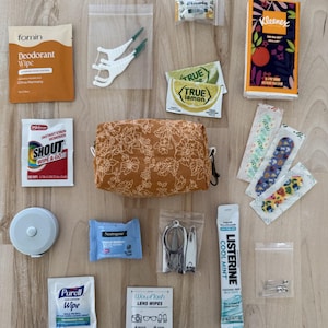 Op de afbeelding: Een mosterdgeel etui met ritssluiting en een wit bloemenpatroon, omringd door diverse reisartikelen voor persoonlijke verzorging, waaronder een Fomin deodorantdoekje, Shout vlekverwijderaar, Kleenex zakdoekjes, True Lemon zakjes en pleisters.
