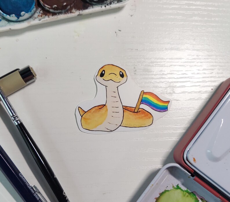 May include: A die-cut sticker featuring a light yellow snake with a tan underbelly and dark eyes. The snake is coiled and holds a small wooden pole with a rainbow-coloured flag. The illustration has a watercolour texture with a thin white border.