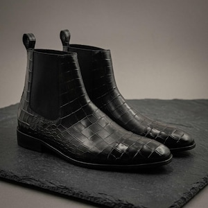 Puede incluir: Un par de botas Chelsea negras con un acabado de cuero texturizado con relieve de cocodrilo. Las botas cuentan con paneles laterales elásticos, punta redondeada, tacón bajo y una trabilla en la parte posterior del tobillo para facilitar el calce.
